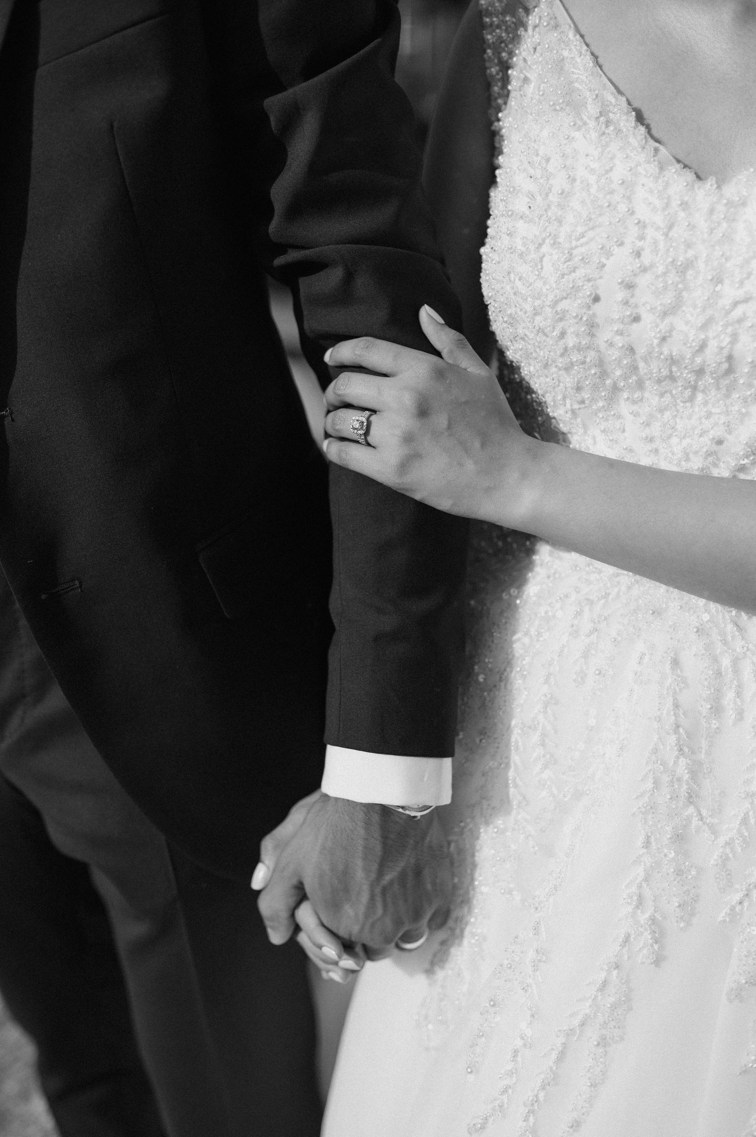 Close-up black and white shot of hands with wedding rings and formal wedding attire.