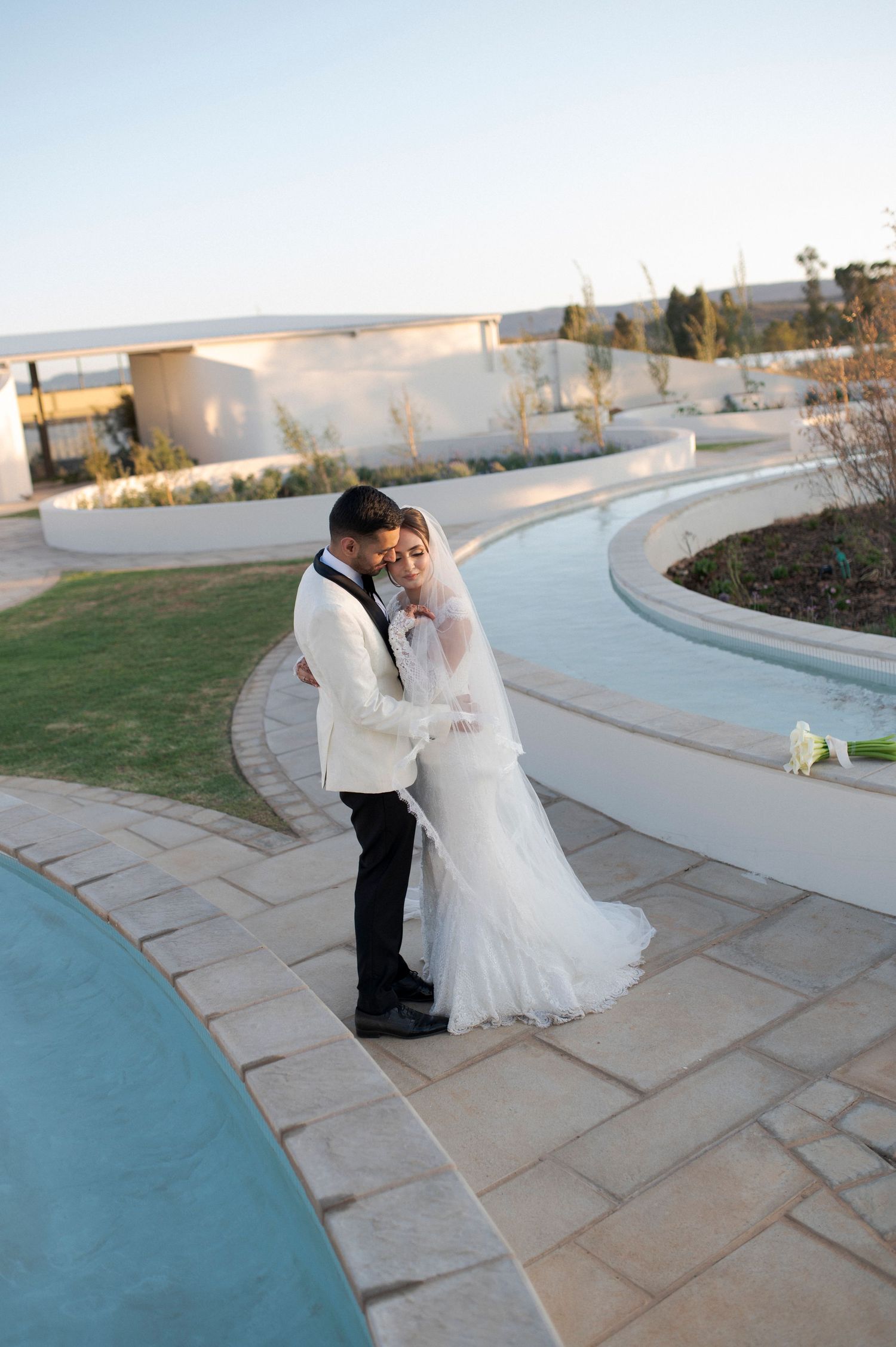 Wedding couple embracing on curved pathway next to pool at sunset outdoor venue.