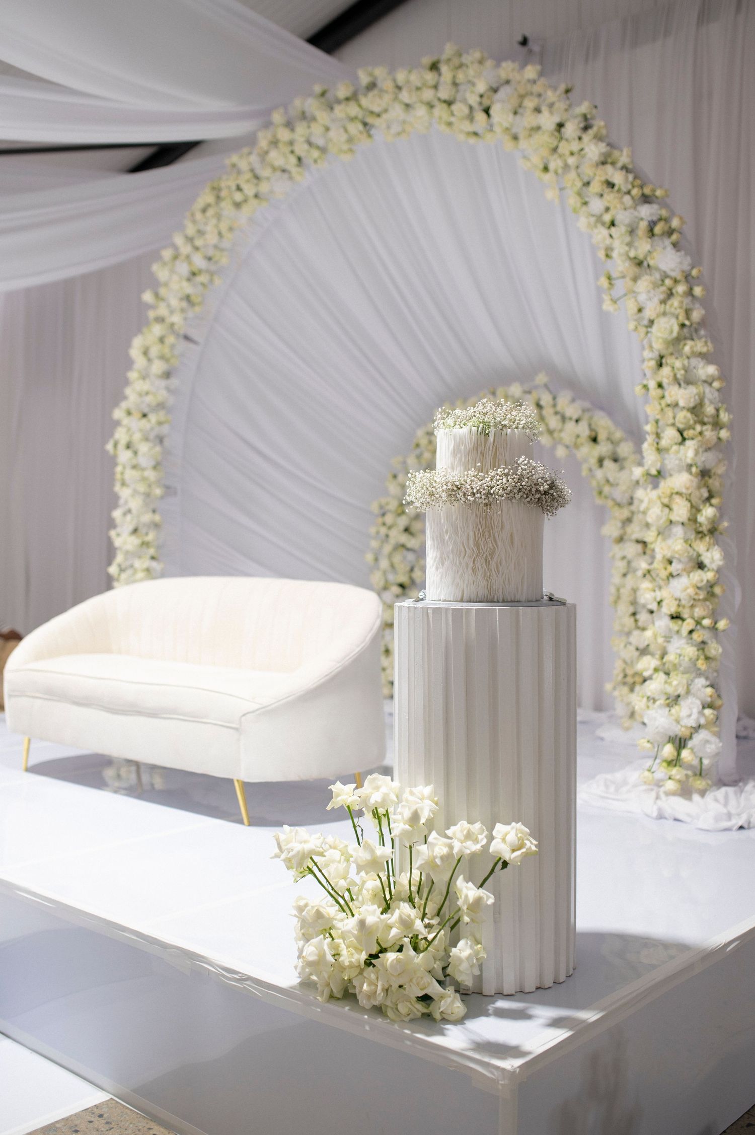 White floral arch with elegant wedding cake display and cream settee on raised platform.