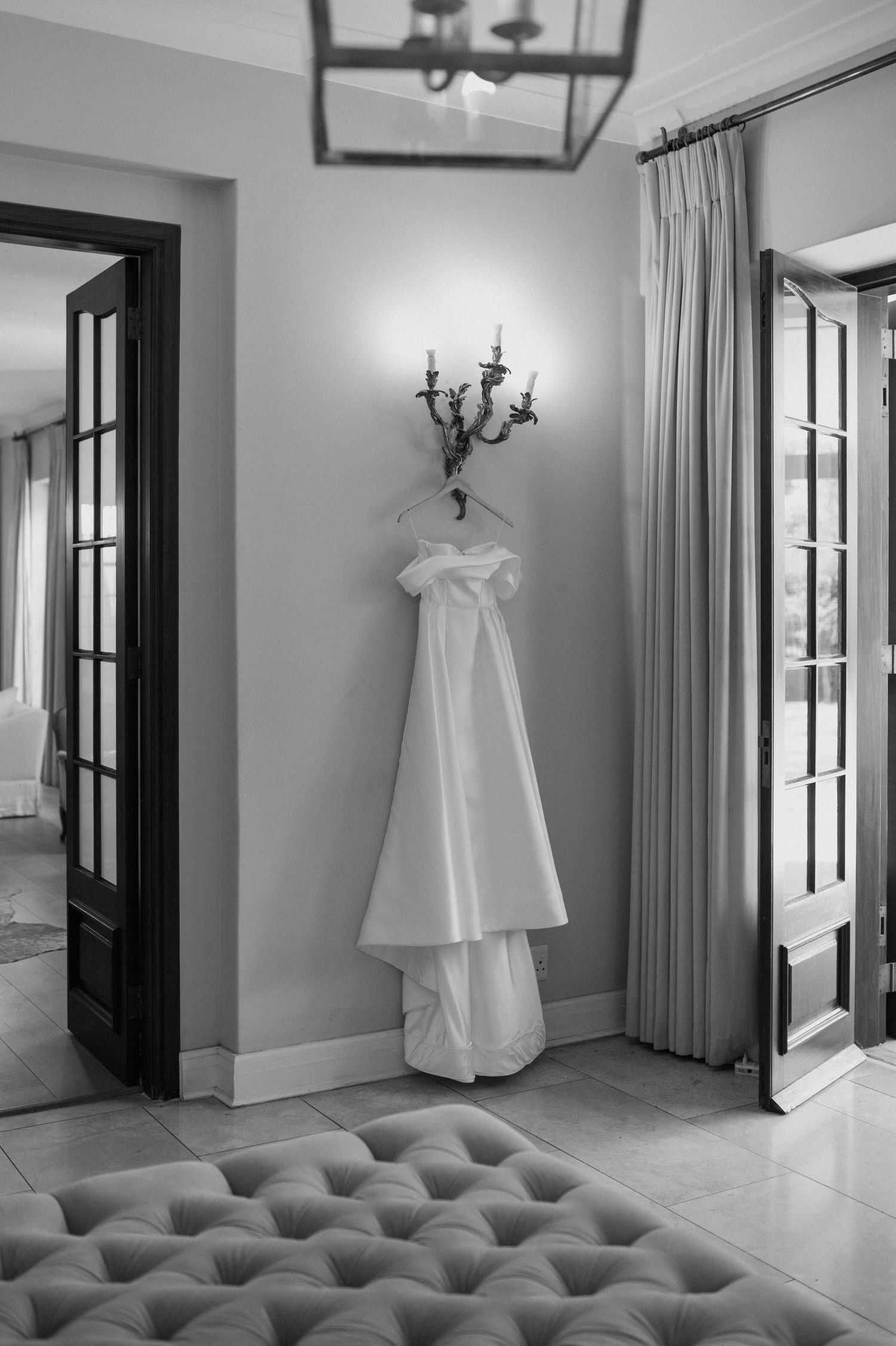 Black and white photograph of a wedding dress hanging on wall beside French doors.
