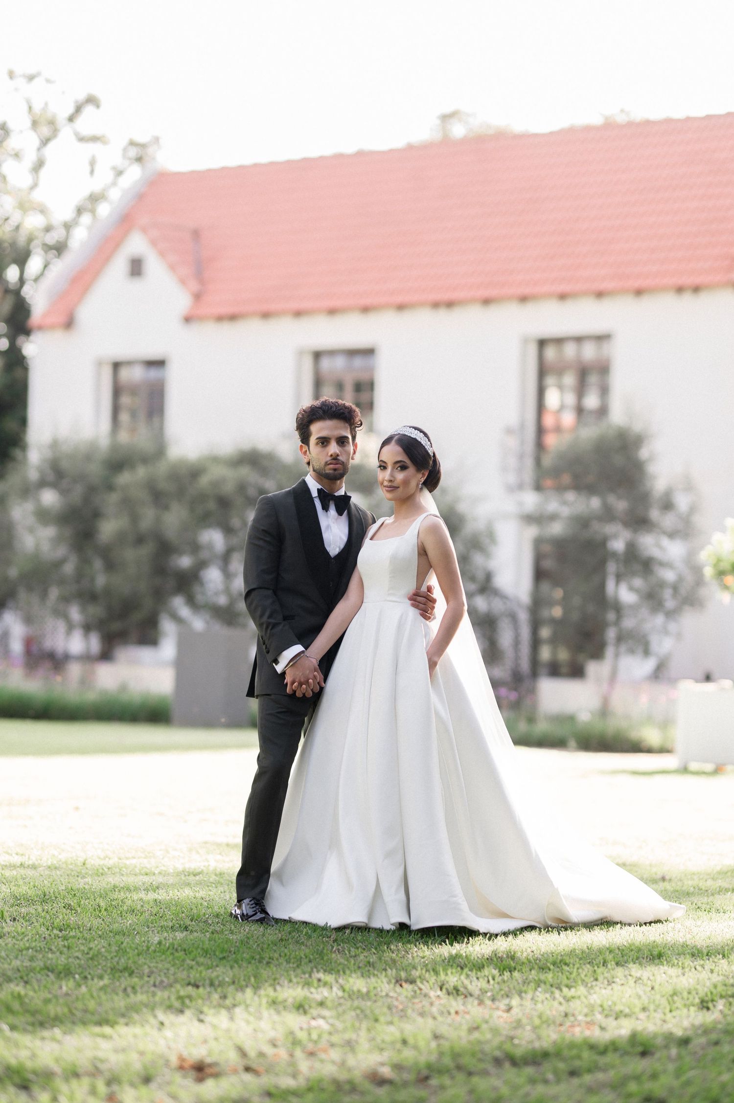 Wedding couple poses in front of white house with red roof in elegant formal attire.