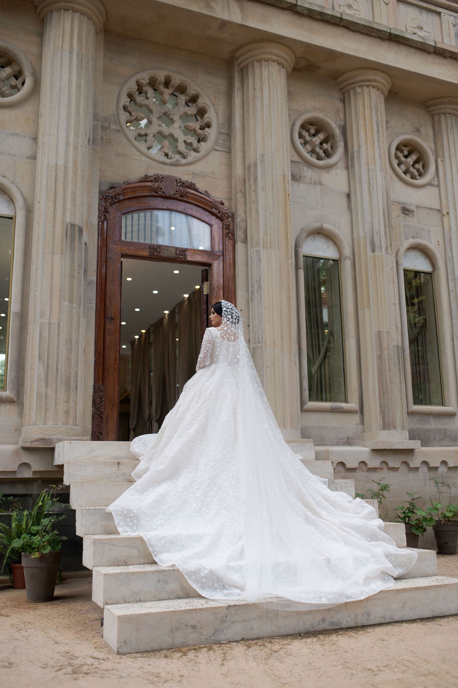 Bride in flowing white wedding dress with long train poses on grand stone staircase.
