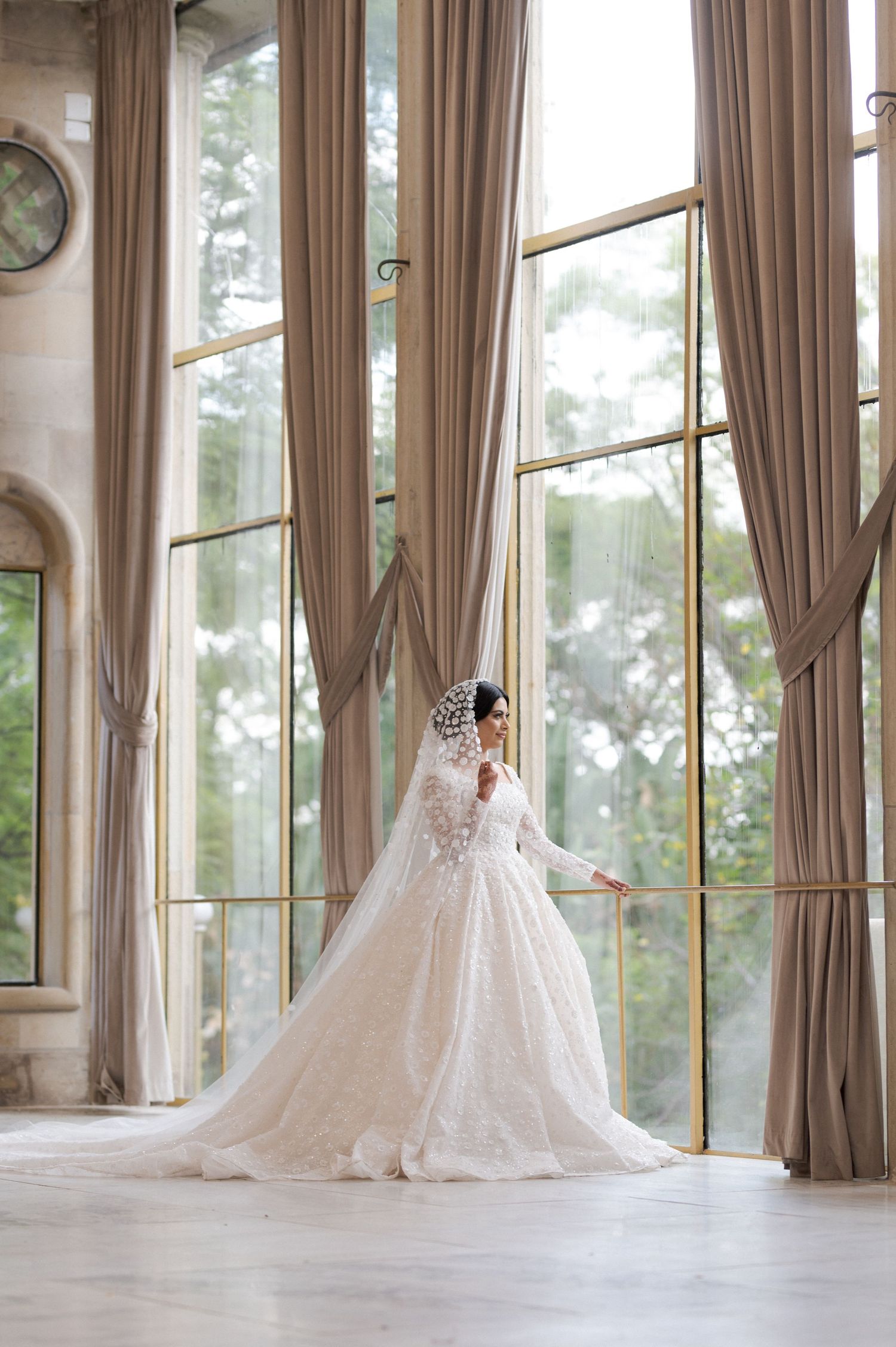 A bride in a flowing white gown poses gracefully by tall windows with floor-length curtains.