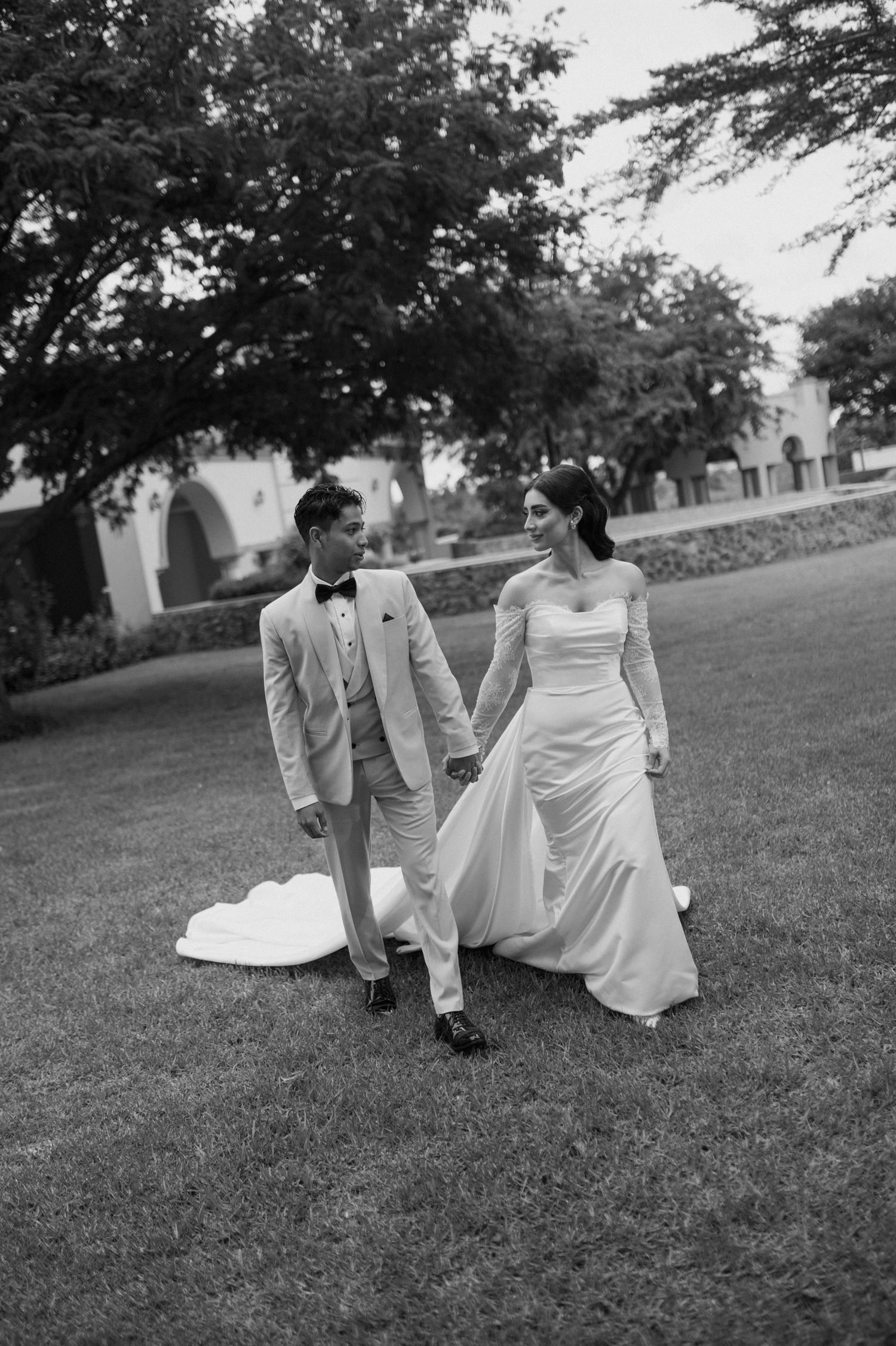 Black and white photo of newlyweds walking together on garden lawn in formal wedding attire.
