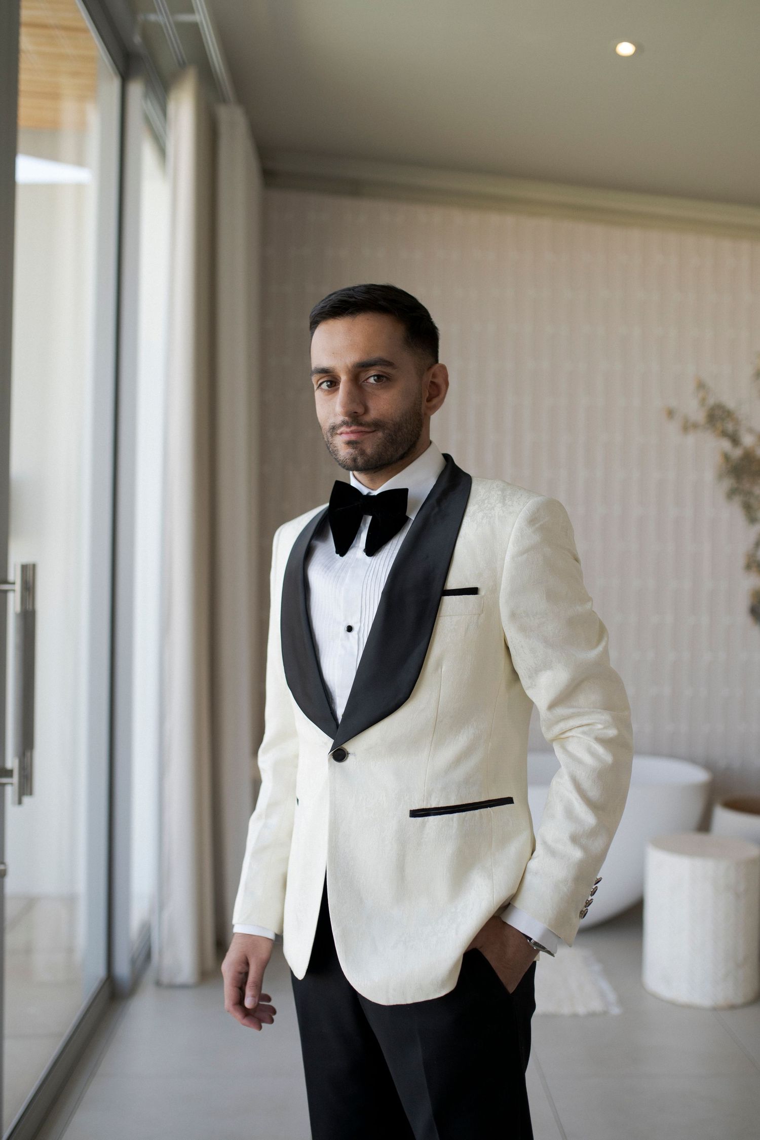 Groom in white and black tuxedo jacket posing by floor-to-ceiling windows.