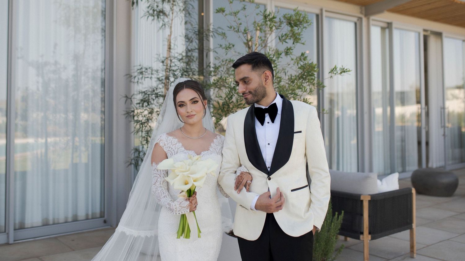 Couple in wedding attire poses on terrace with glass windows and greenery.