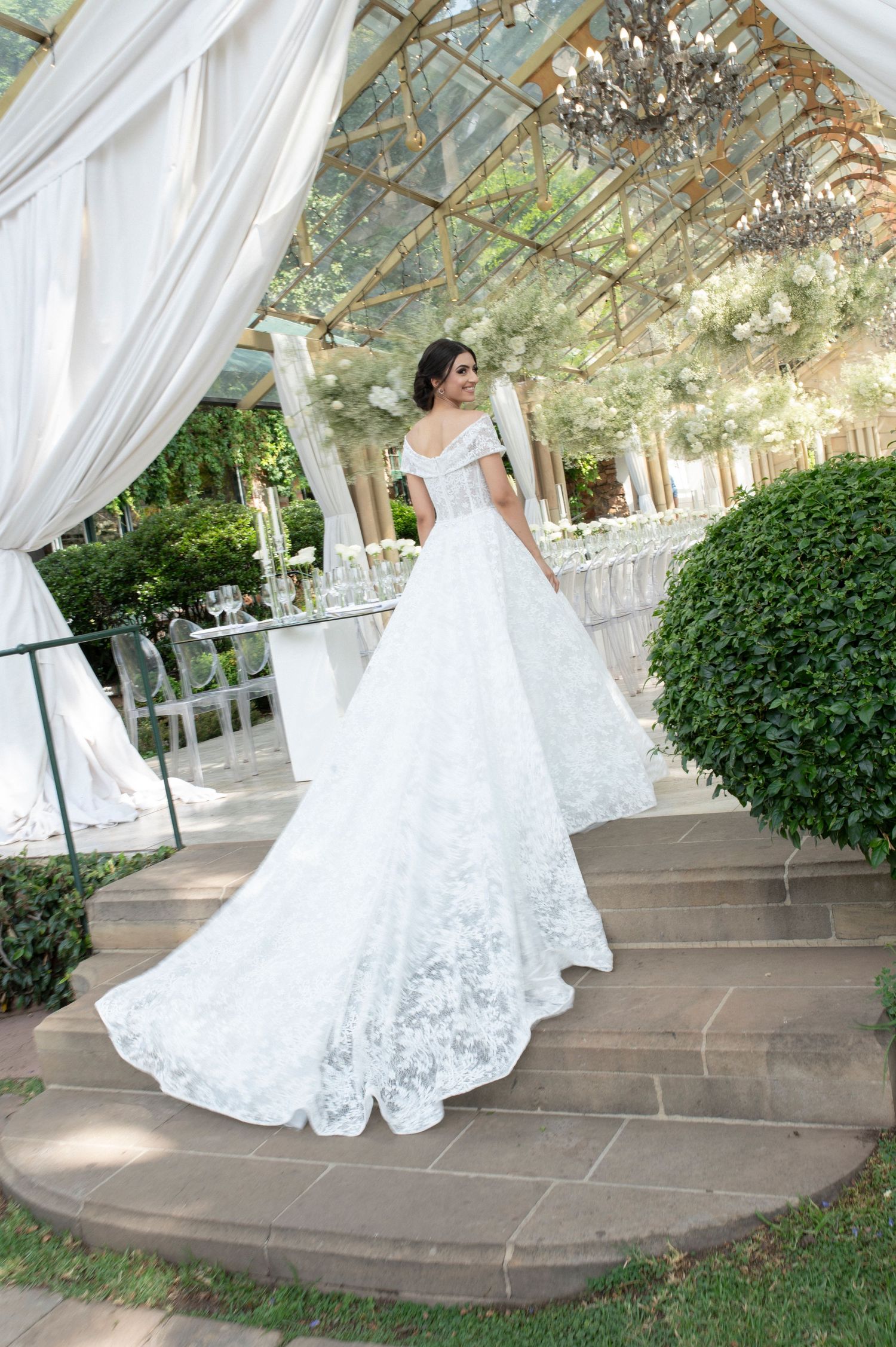 Bride in off-shoulder ballgown stands on garden steps under glass conservatory ceiling.