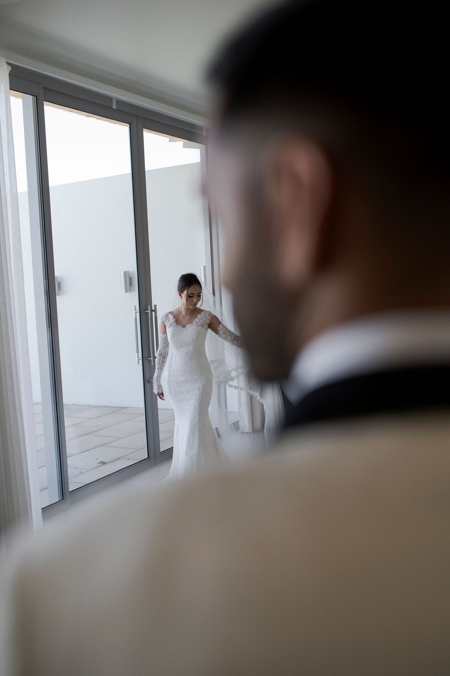 Through a glass door, a bride in a white lace gown is seen while a groom looks on in silhouette.