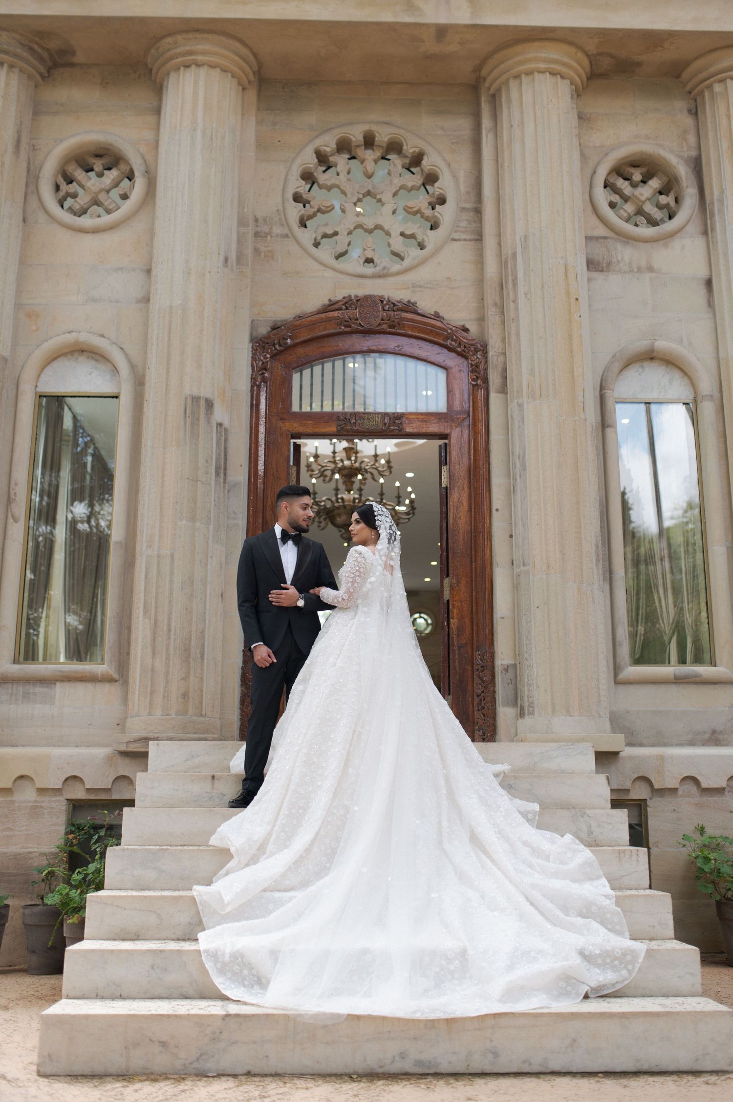 Couple poses on grand marble steps in front of elegant doorway, bride wearing stunning long-train white gown.