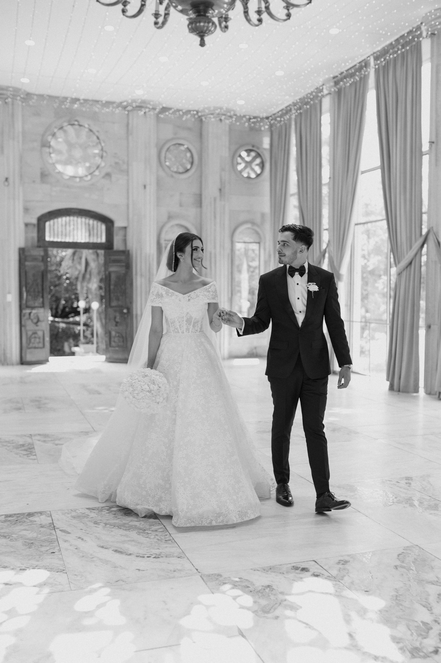 A couple shares an elegant moment in a grand ballroom with ornate marble floors and chandeliers.