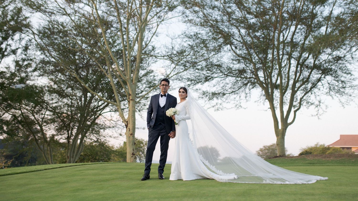 Wedding couple poses on golf course with dramatic flowing veil and trees in background.