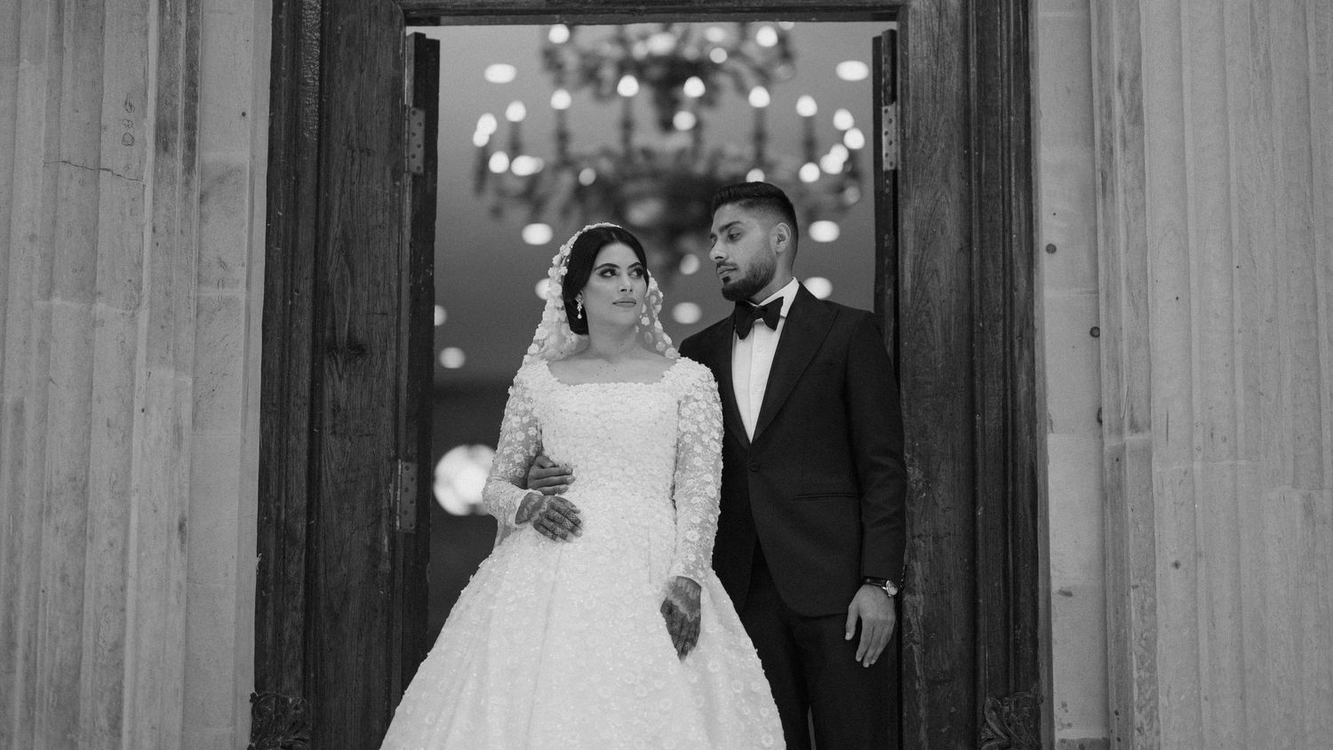 Black and white wedding portrait showing a couple embracing in doorway with twinkling lights in background.