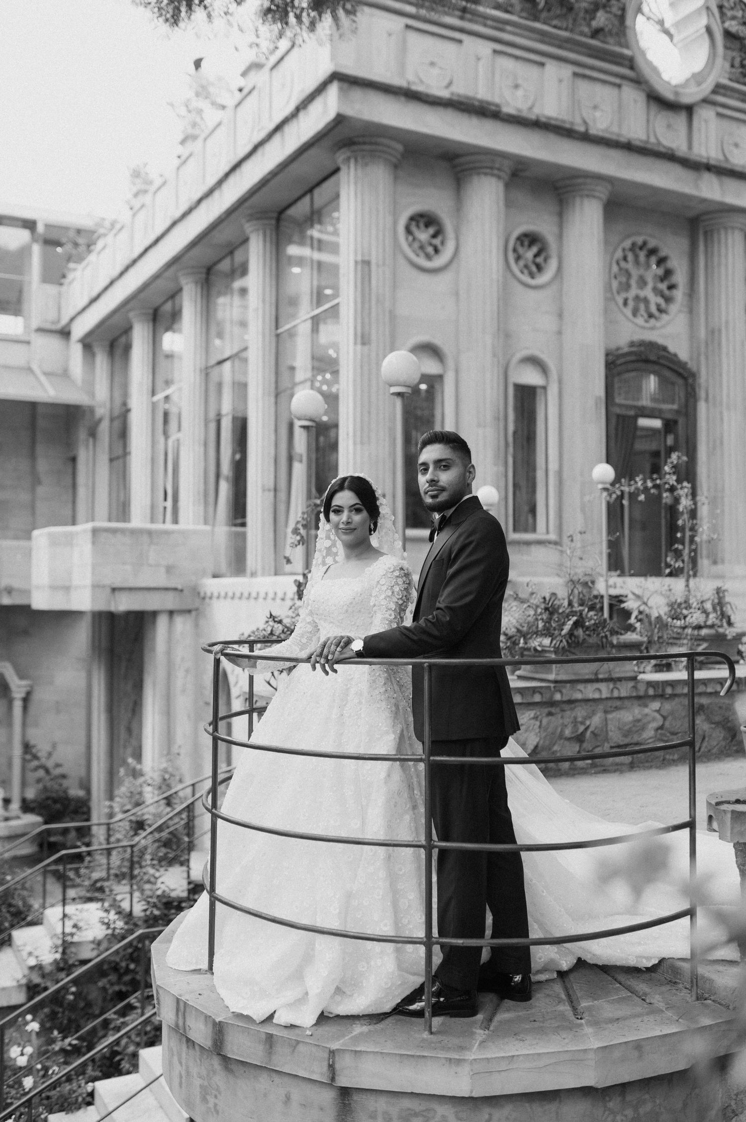 Wedding couple stands on vintage balcony with ornate architectural details in black and white photo.