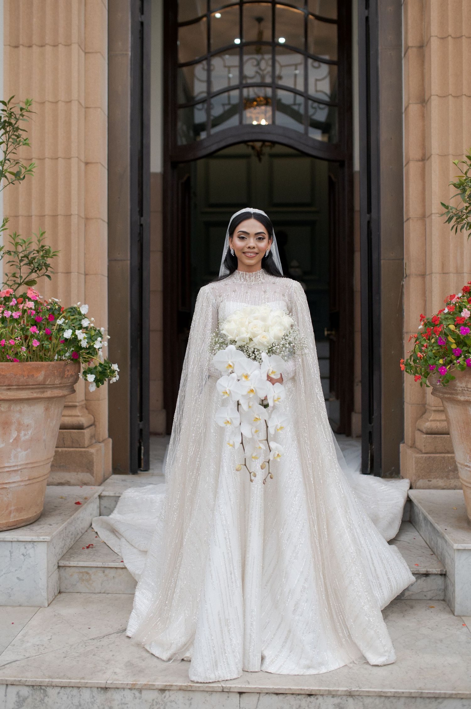 A bride in a high-neck wedding dress poses on stone steps with potted flowers and classical architecture.