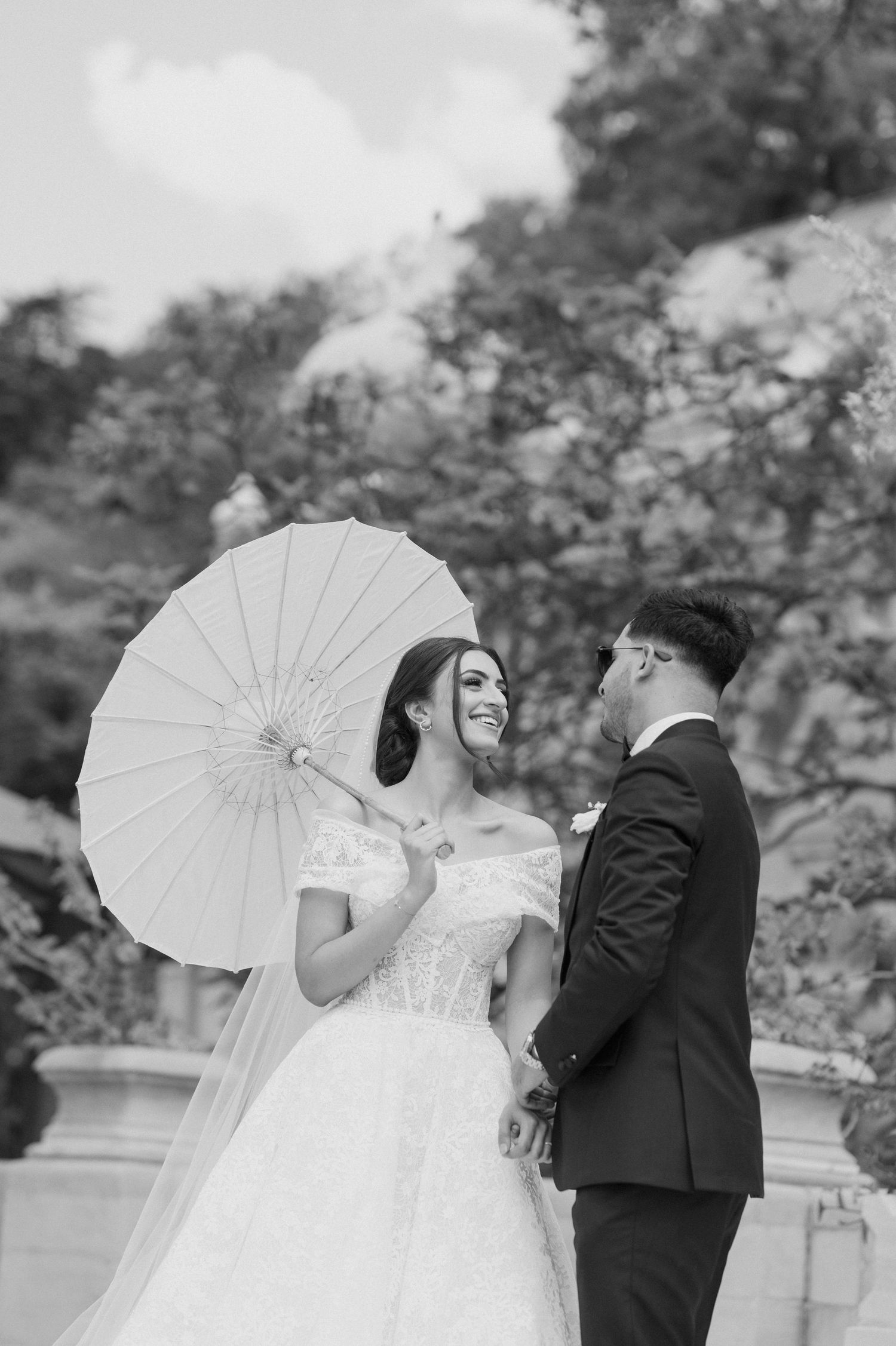 A black and white wedding photo featuring a couple with a decorative white parasol outdoors.