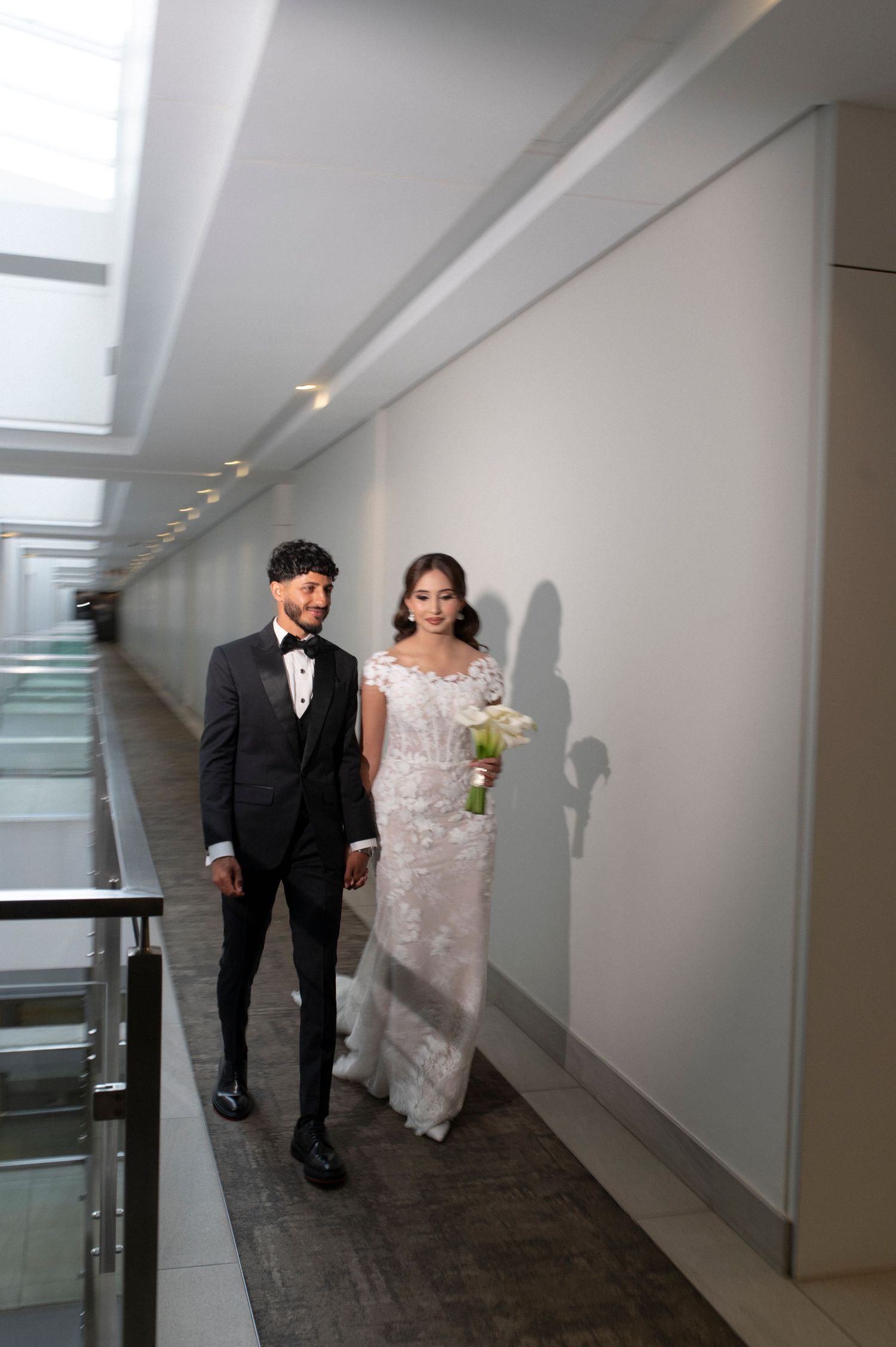 Wedding couple walking together in a modern hallway with floor-to-ceiling windows.