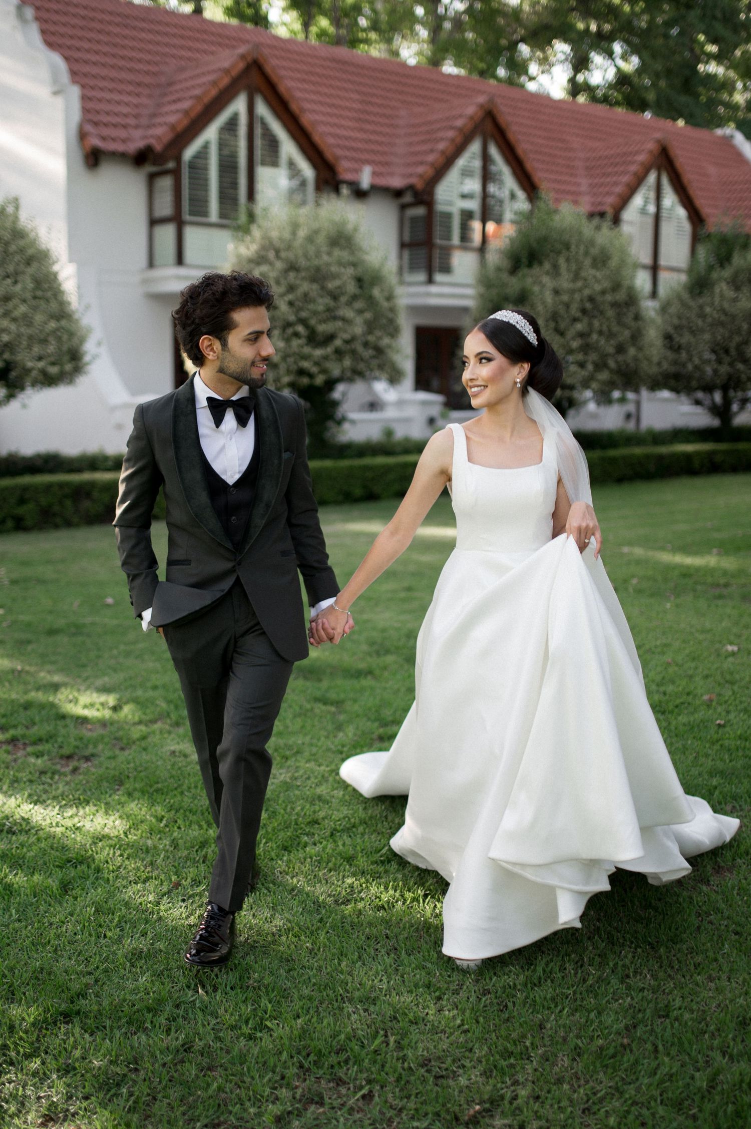 Bride and groom walking hand in hand on garden lawn with Tudor-style buildings in background.