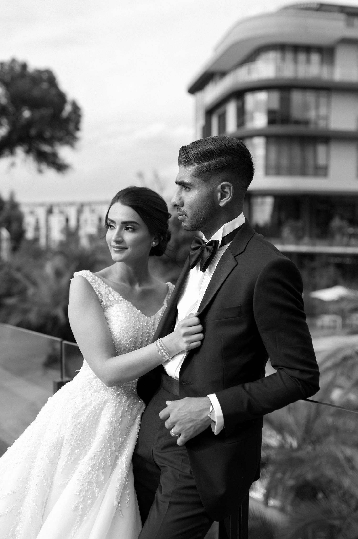 An elegant wedding couple poses together on a balcony with modern architecture in the background.