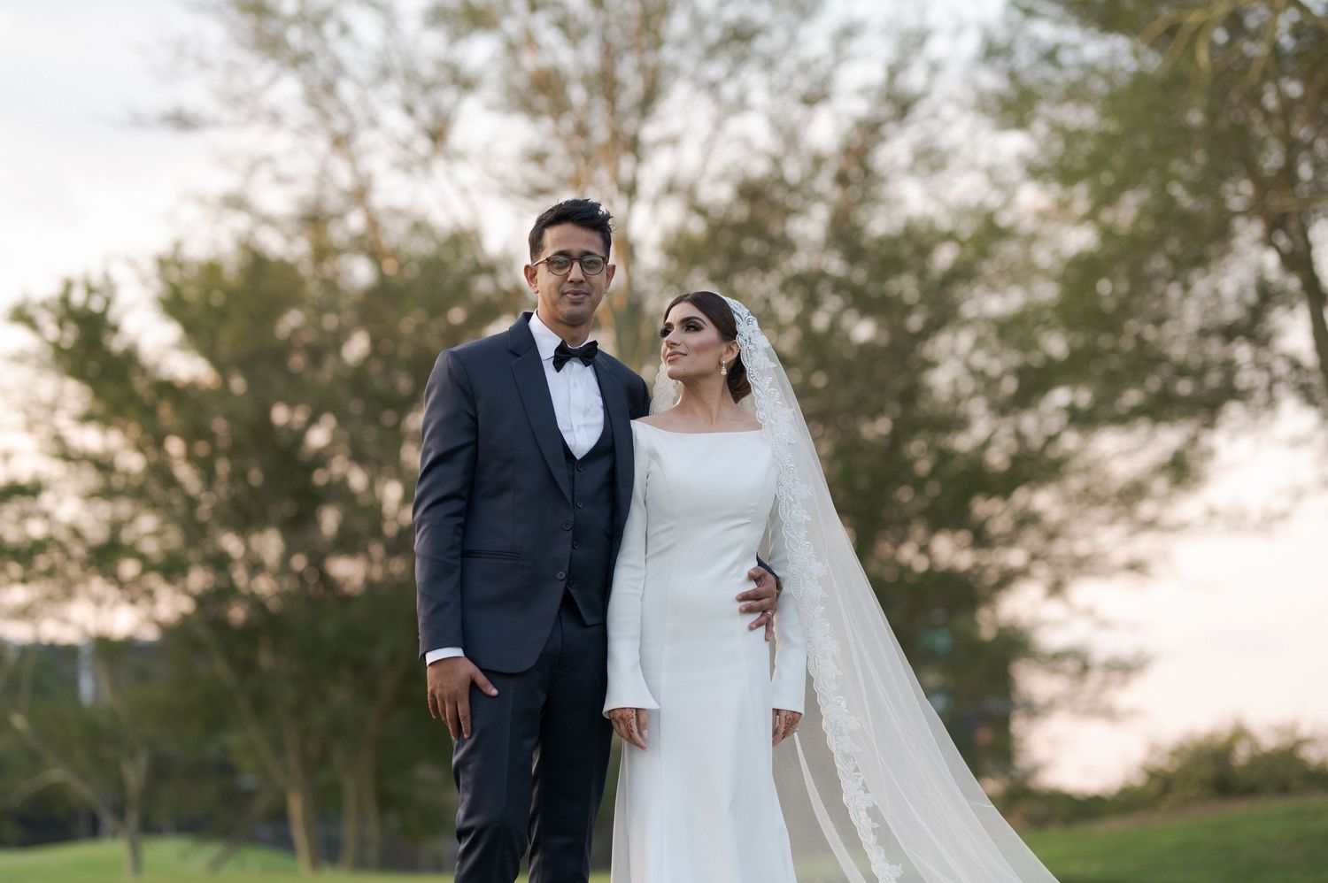 Newlywed couple standing outdoors at sunset with bride wearing long-sleeved gown and flowing veil.