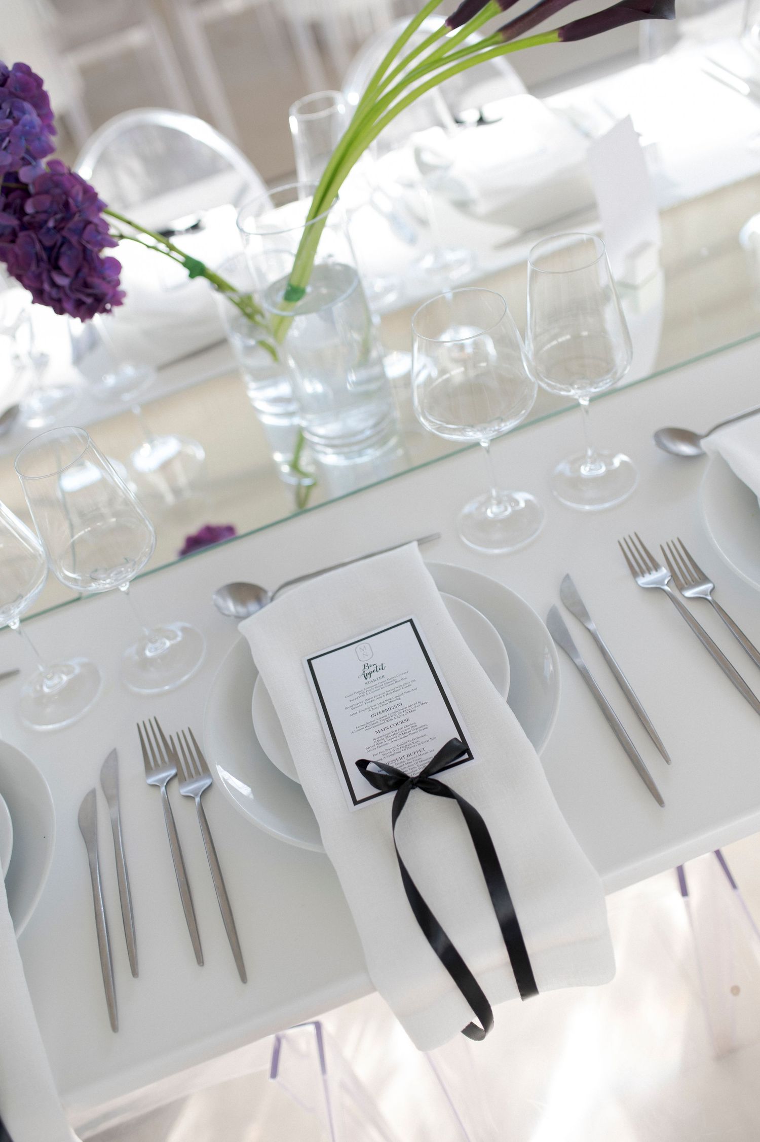 Elegant white place setting with menu card tied with black ribbon and purple flowers in glass vase.