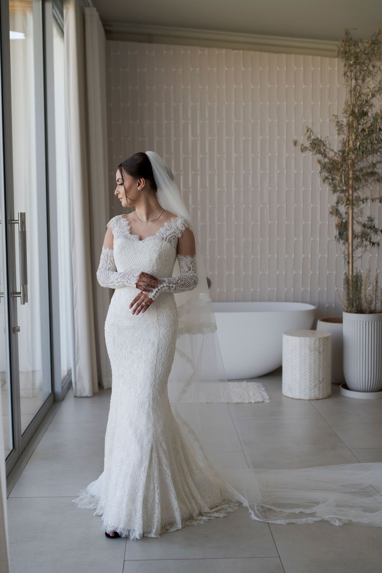 Bride in long-sleeve lace wedding dress posing by window with modern bathroom decor.