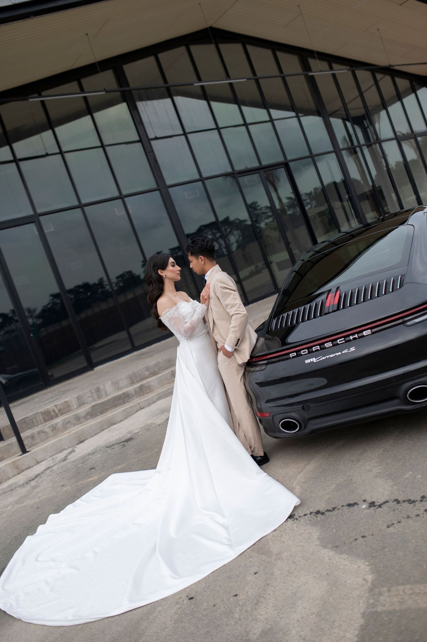 Wedding couple poses by modern building with luxury car in background.