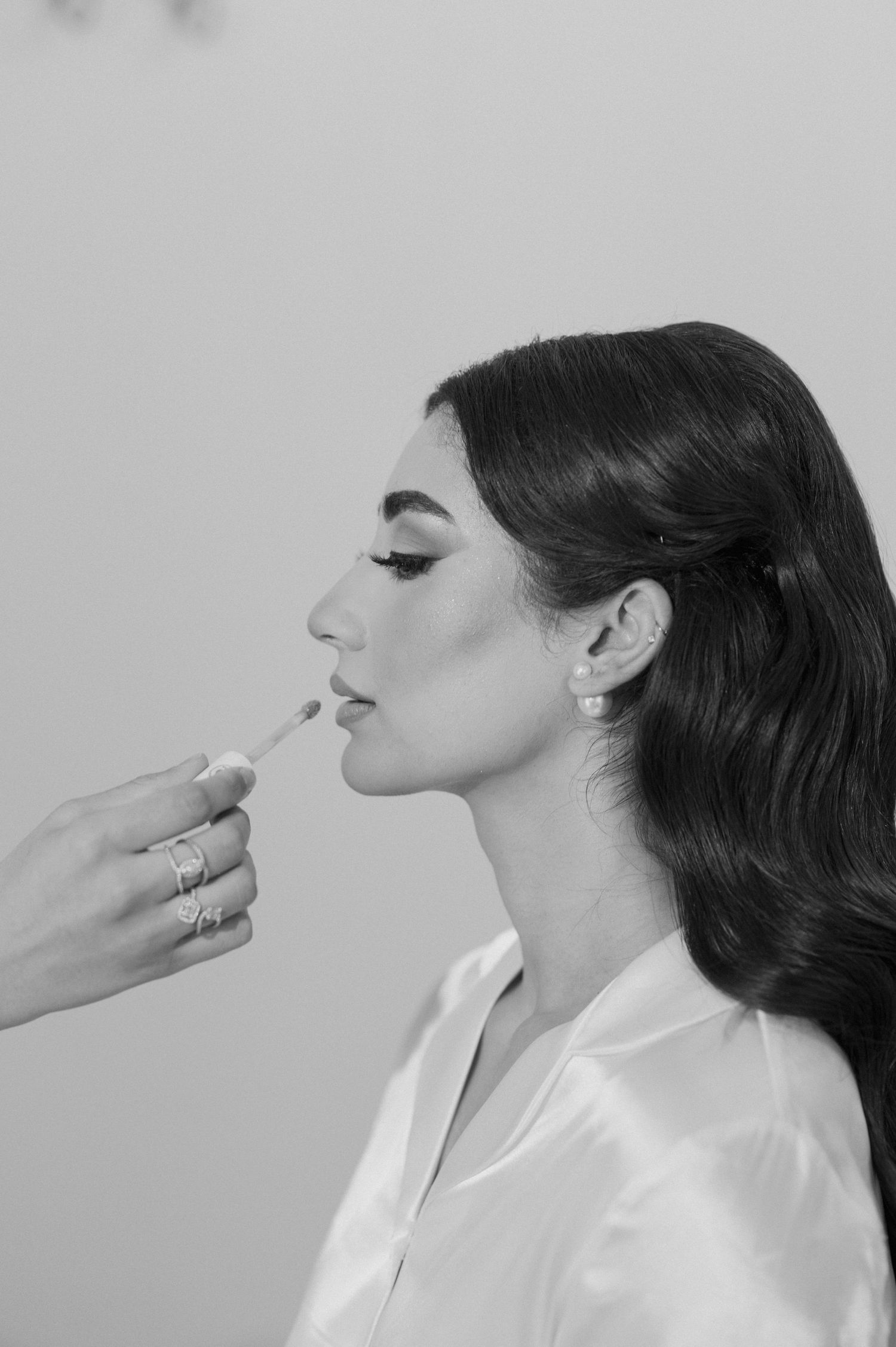 Black and white bridal portrait showing makeup application with dark wavy hair and pearl earrings.
