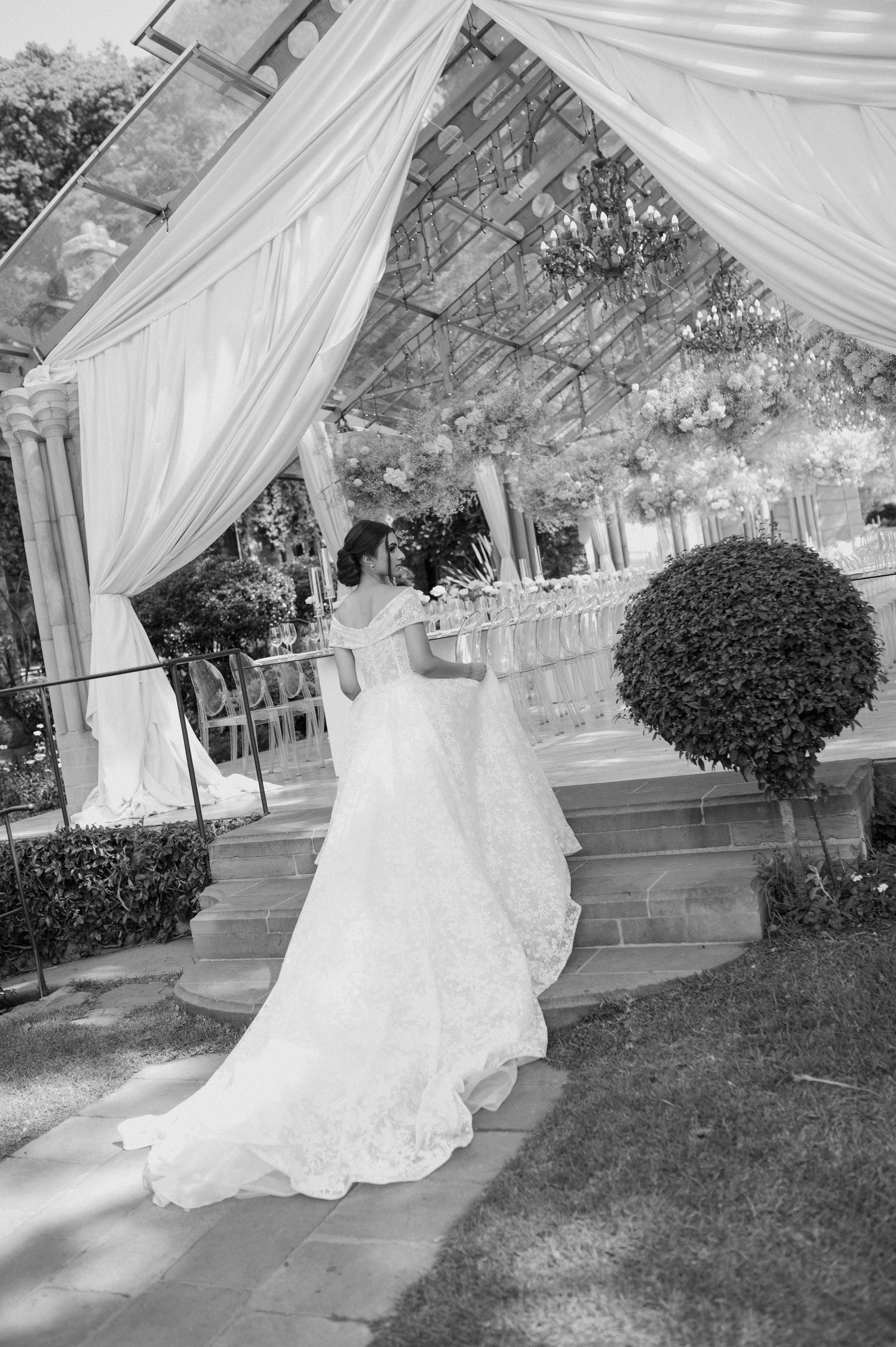 An elegant bride in a flowing white gown stands beneath a draped outdoor wedding canopy.