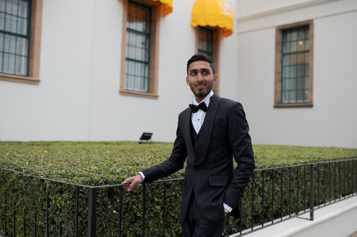A groom in a black tuxedo poses beside a wrought iron railing with yellow awnings in background.