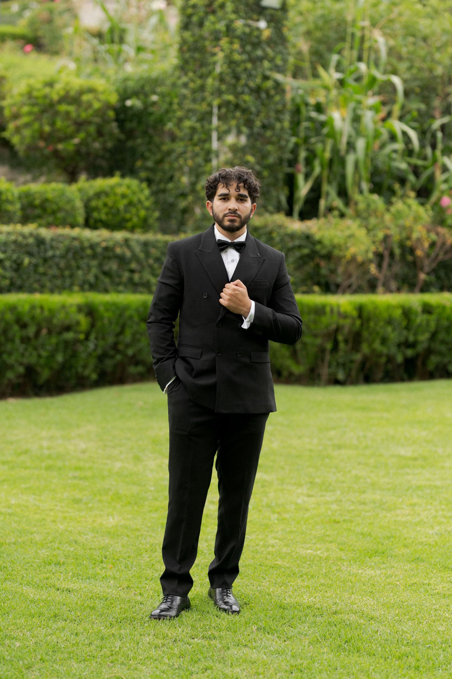 A groom in a black tuxedo and bow tie poses on manicured lawn with garden backdrop.