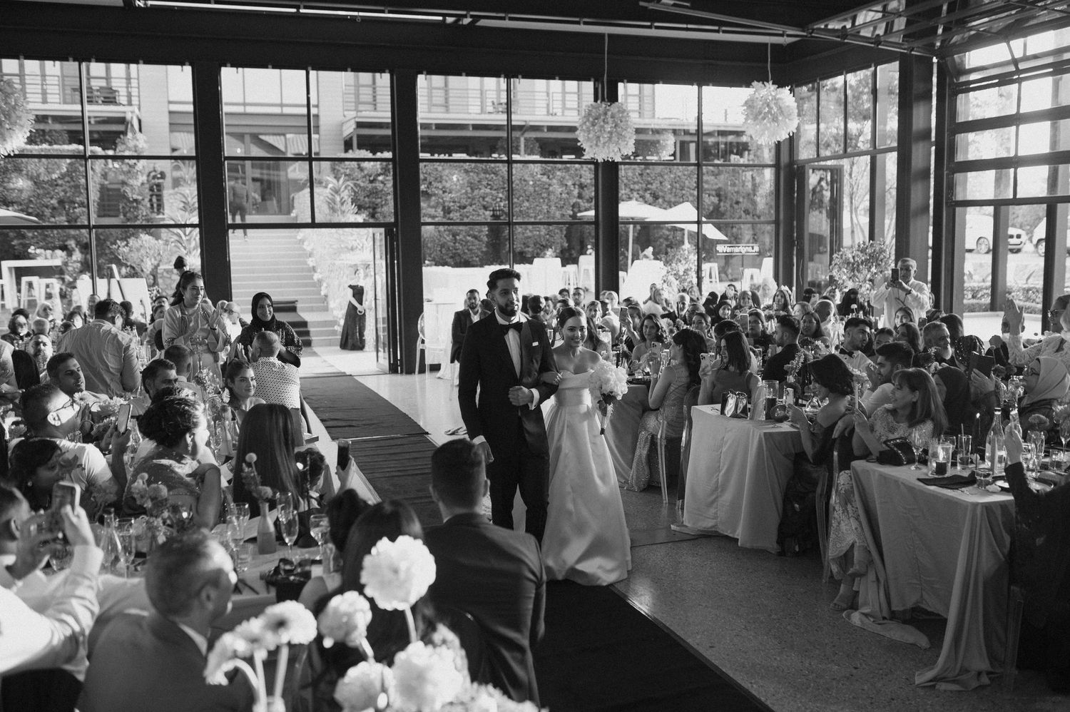 Black and white photograph of an indoor wedding ceremony with guests seated at round tables.