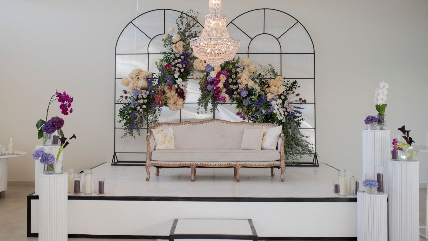 Wedding venue setup with floral arch, crystal chandelier and elegant white seating.