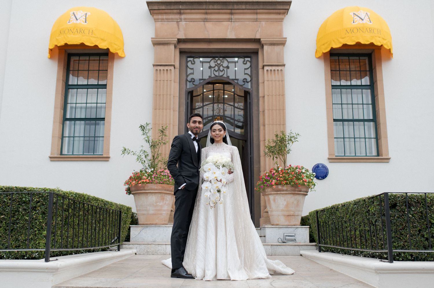 A wedding couple poses in front of an ornate doorway with yellow awnings and manicured hedges.