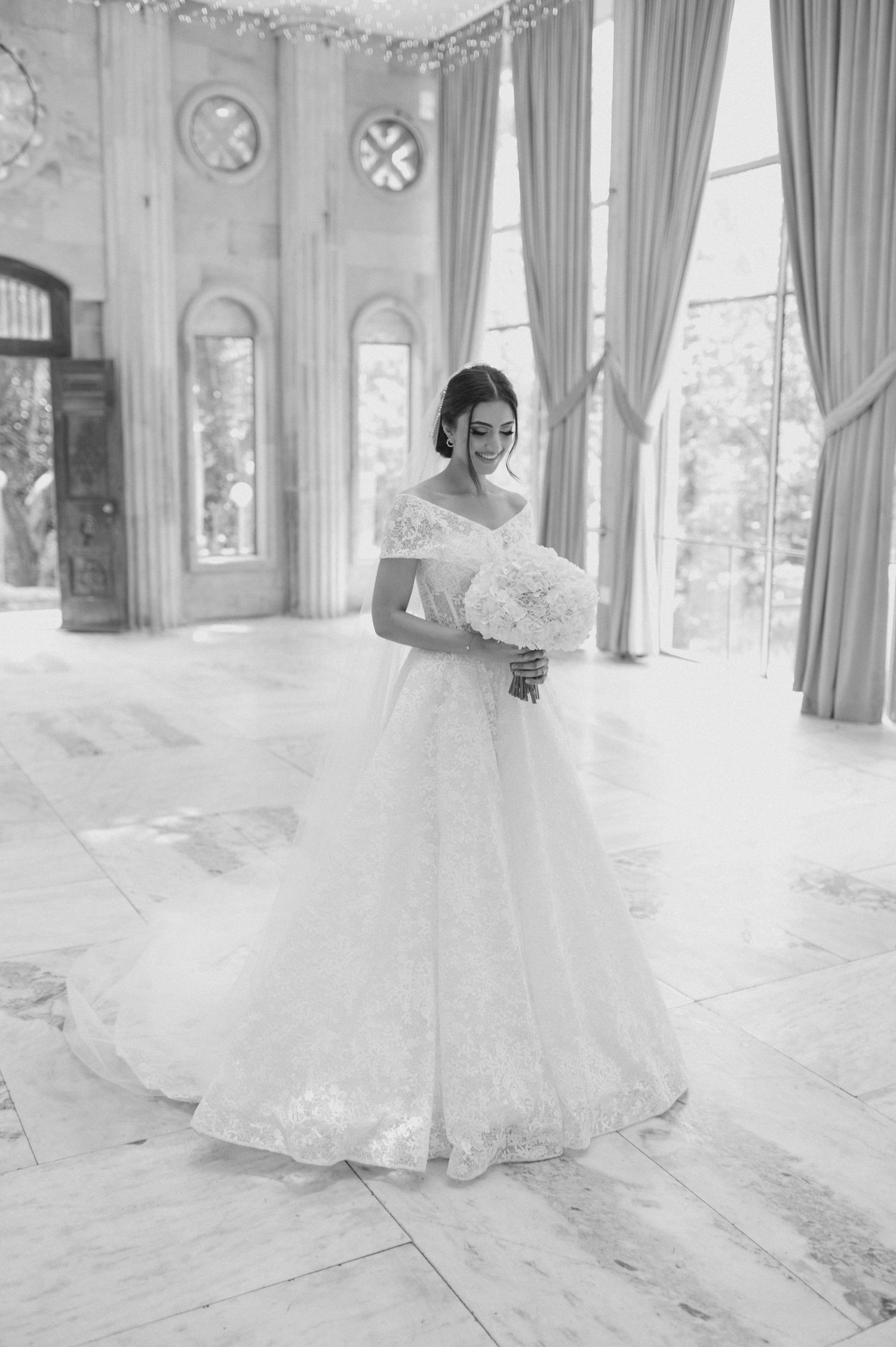 Elegant bride in off-shoulder lace wedding gown holds white bouquet in grand marble room with tall windows.