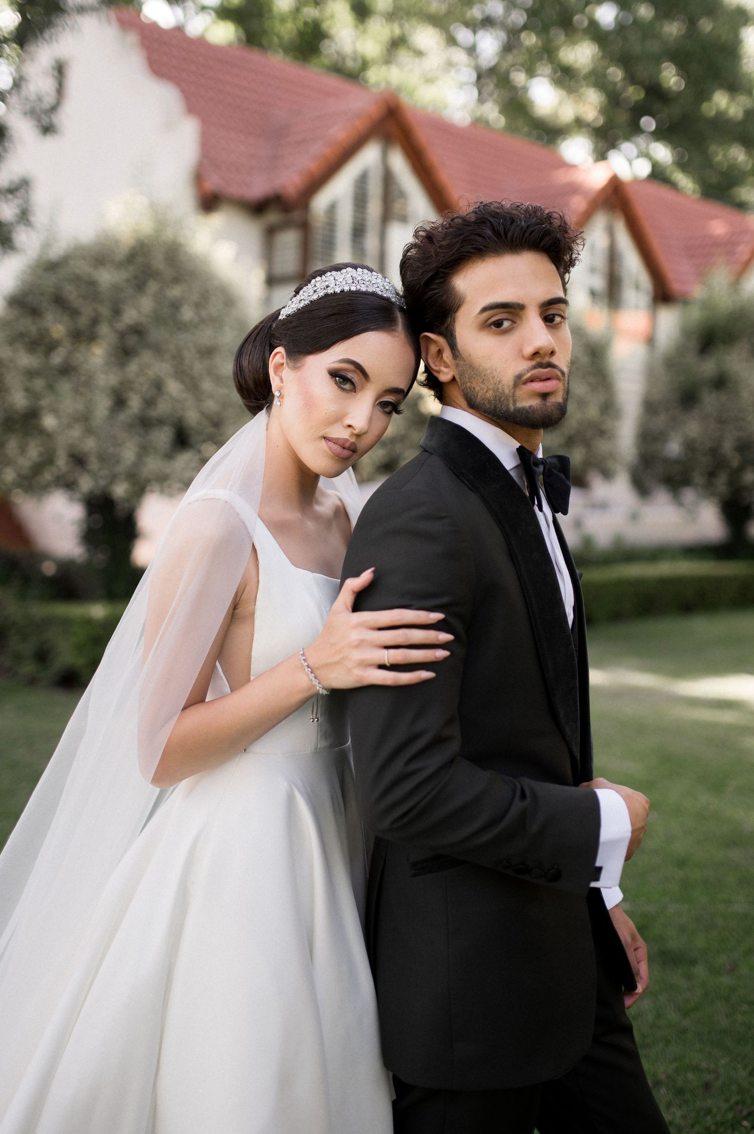 A romantic wedding portrait of a couple in formal attire posing in front of Tudor-style houses in a garden setting.