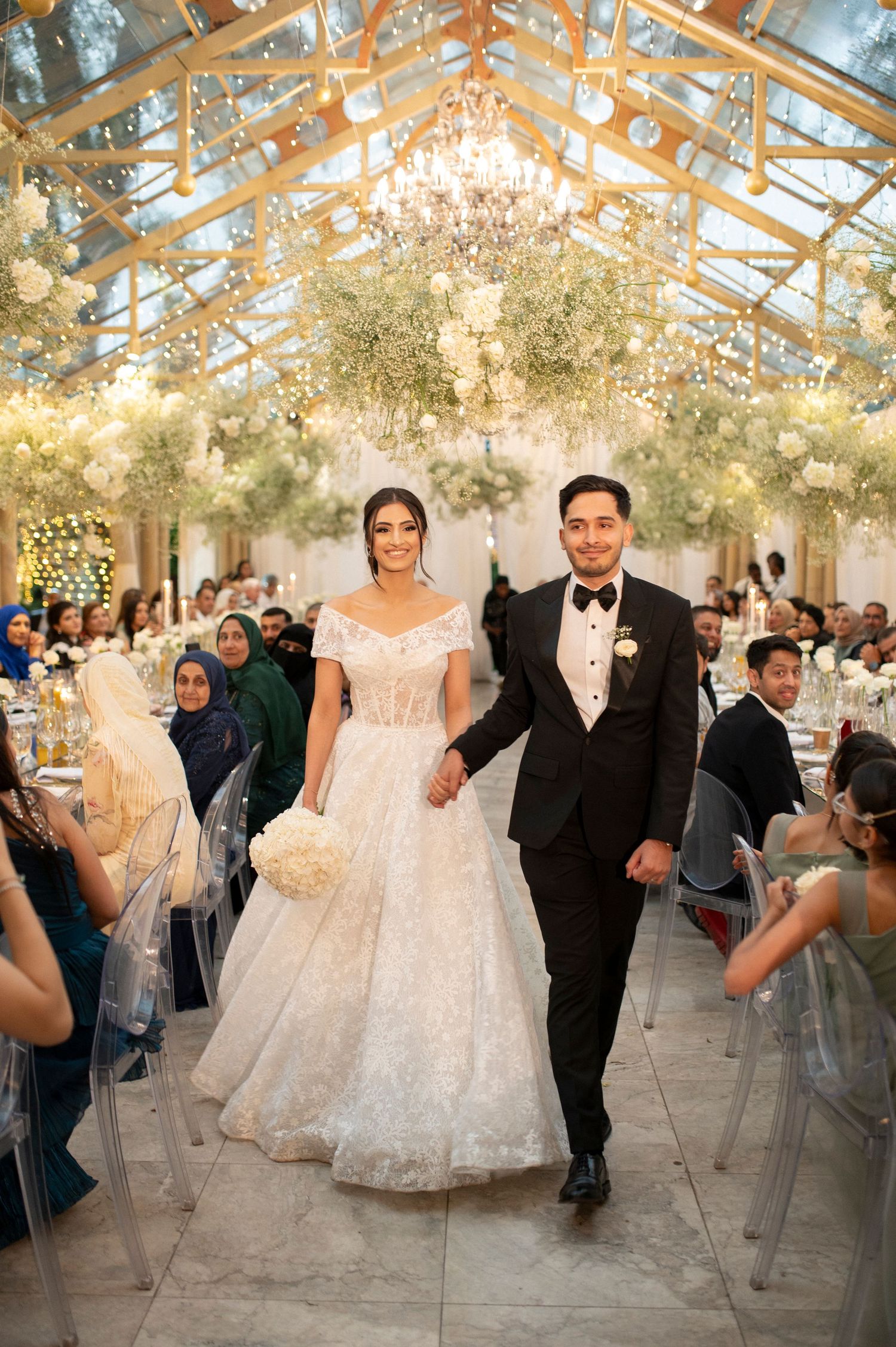 Wedding reception in a glass-ceiling venue decorated with ethereal floral installations and chandeliers.