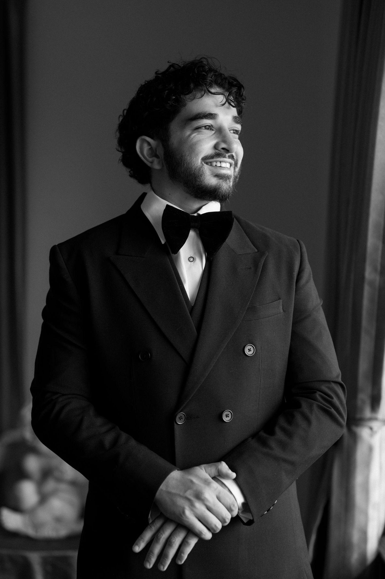 Classic black and white portrait of groom in tuxedo smiling by window.
