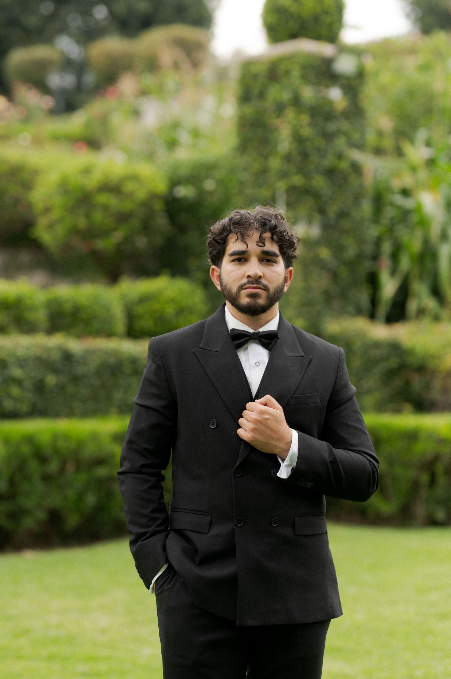 A groom adjusts his bow tie while standing on grass with landscaped hedges behind him.