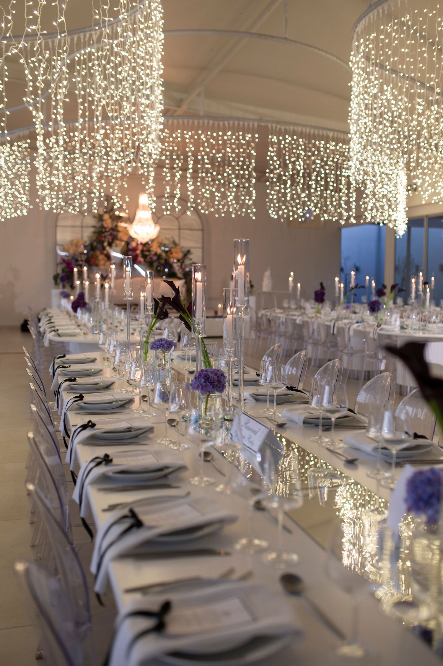 Elegant wedding reception table setting with cascading fairy light curtains and purple floral accents.