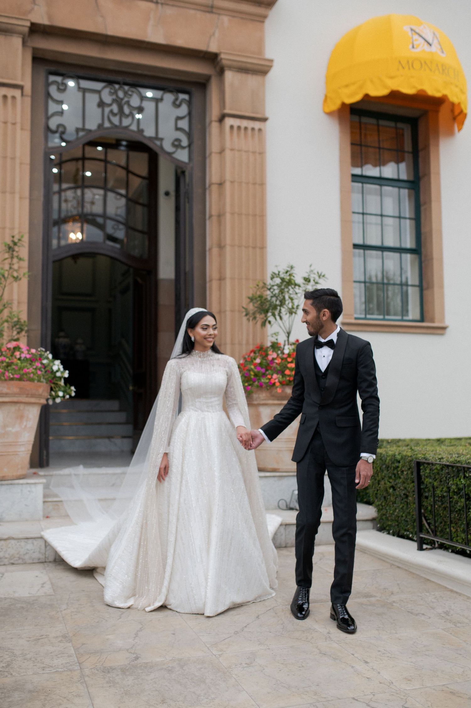 A couple walks hand in hand outside a building with yellow awnings, bride in white and groom in black tuxedo.