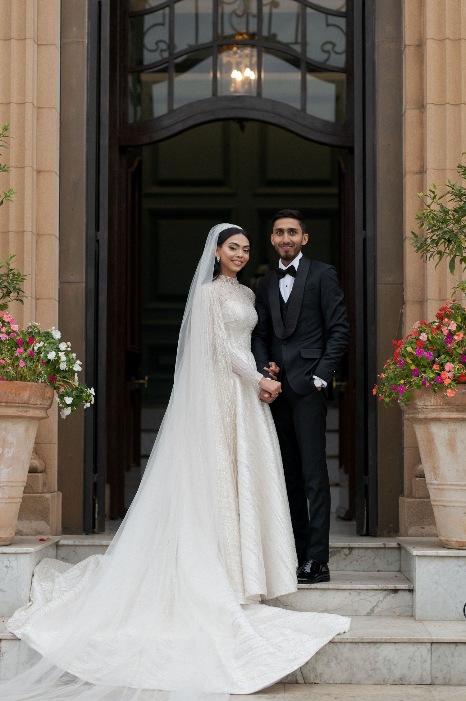 A newlywed couple stands on stone steps before a grand entrance with floral decorations.