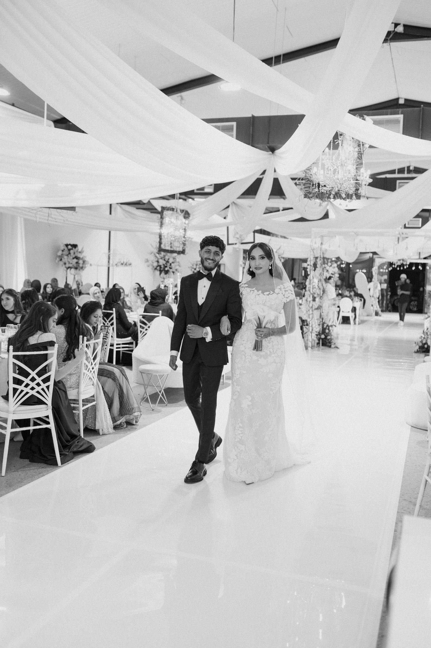 Wedding ceremony processional in a white draped venue with guests seated along the aisle.