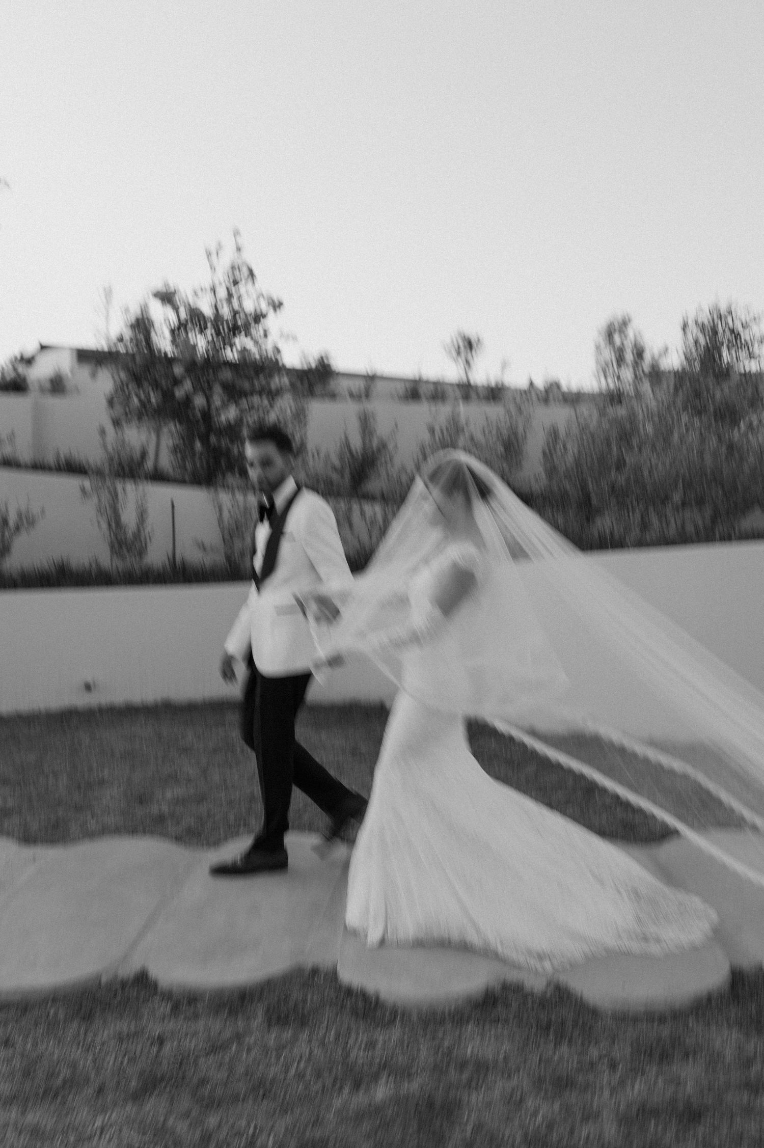 A black and white photo of newlyweds walking along an outdoor pathway with flowing veil.