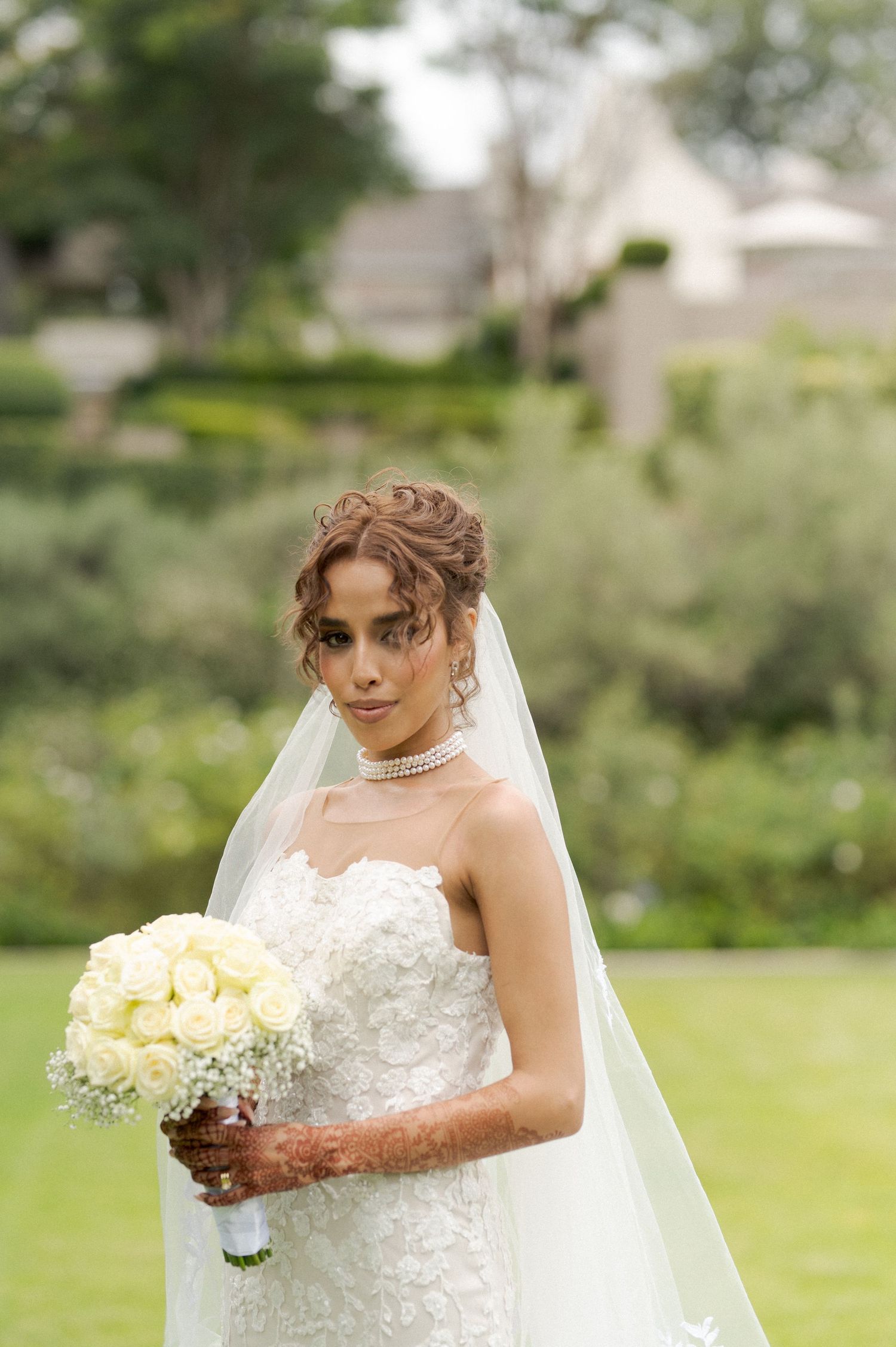 A bride in a white lace gown holds a cream bouquet while wearing a flowing veil in an outdoor garden setting.