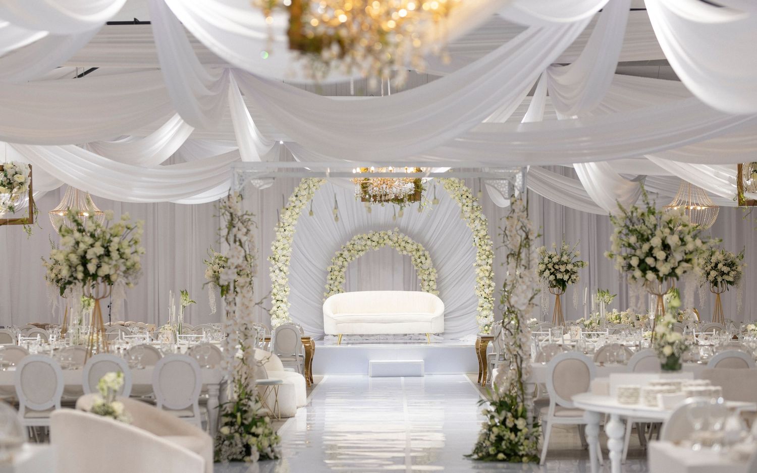 Luxurious white wedding venue with floral arch, draped ceilings and elegant table settings.