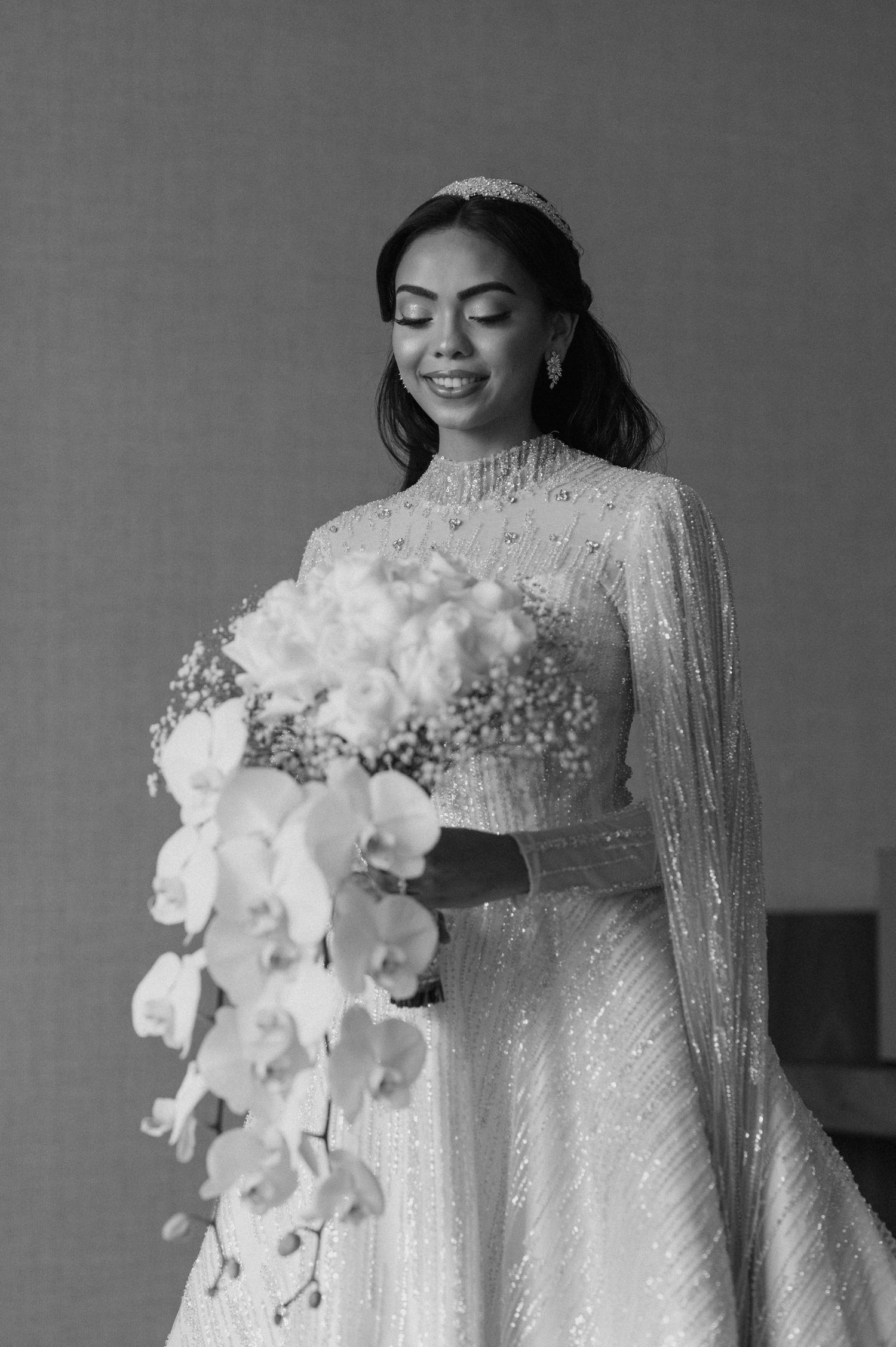 Bride in high-neck beaded gown holds cascading white orchid bouquet in black and white portrait.