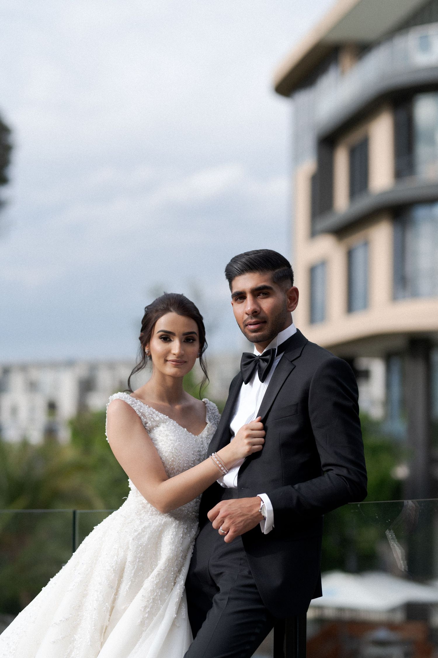 A wedding couple poses elegantly against a modern building backdrop in formal attire.
