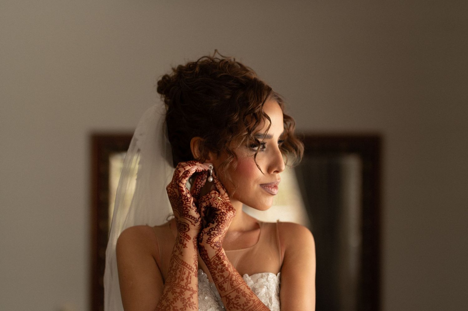 A bride with henna decorated hands touches her earring in soft window light.