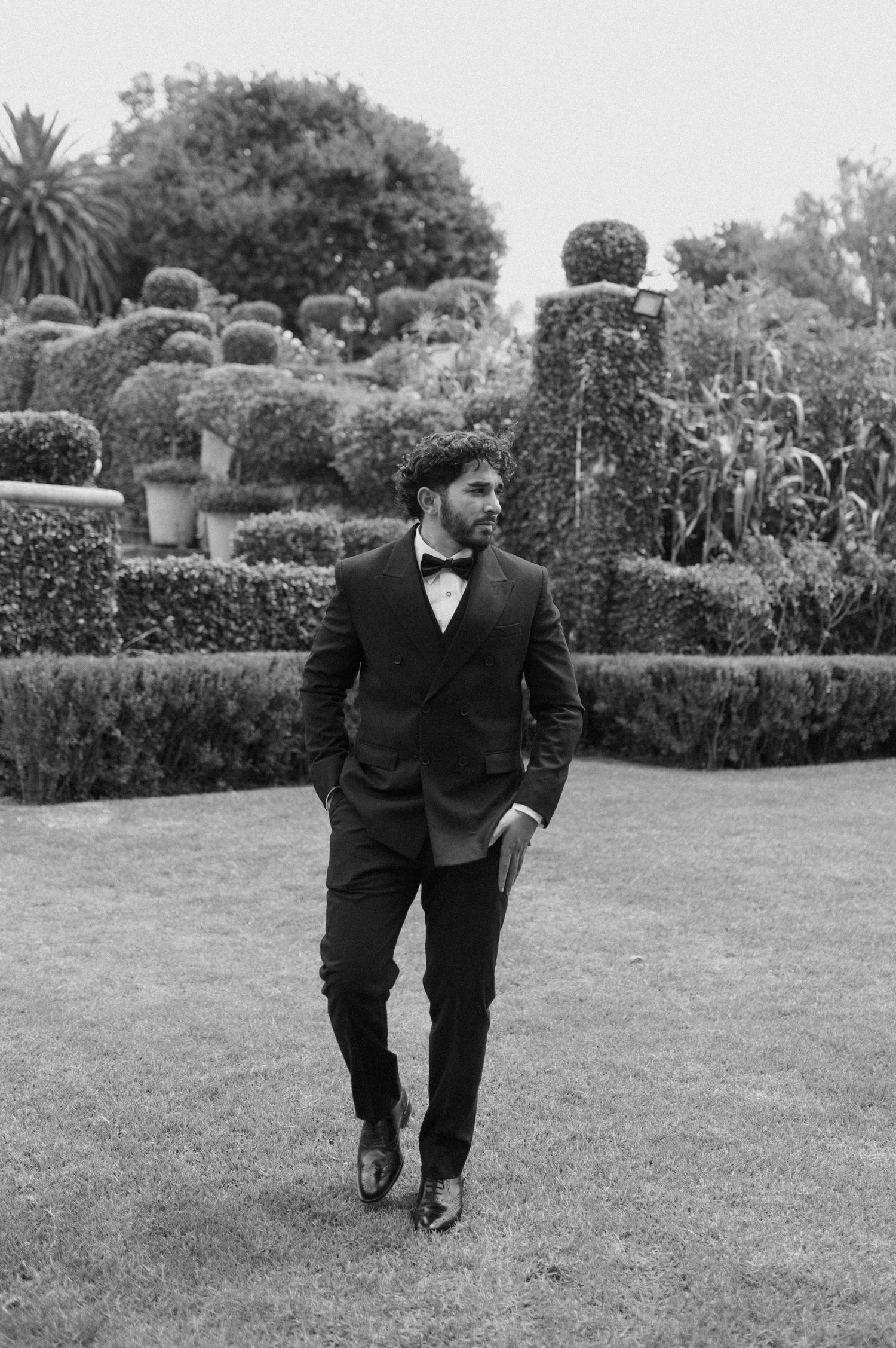 Black and white photo of a groom in a tuxedo walking through a manicured garden.