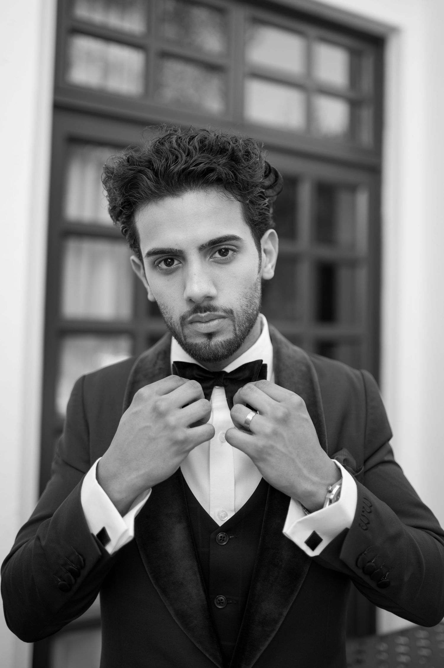 Groom adjusts bowtie while wearing classic black tuxedo in elegant black and white portrait.